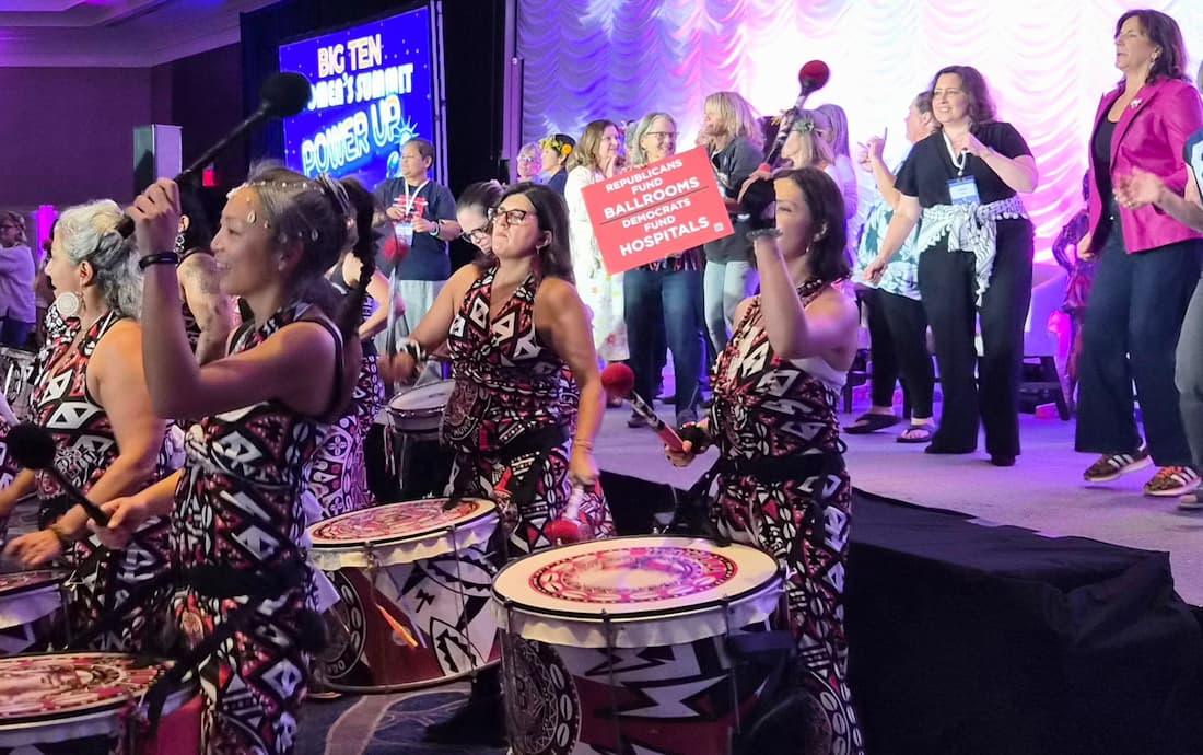 Drum line performing at the Big Ten Women's Summit with a Republicans Fund Ballrooms, Democrats Fund Hospitals sign held up in the crowd