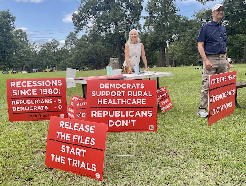 Volunteers with a table of Recessions, Healthcare, Groceries, Release the Files, and Vote This Year signs at an outdoor event