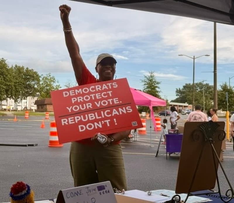 Five people holding Democrats Protect Your Vote, Healthcare, Lower Prices, Trump's War, and Jobs Created signs on a lawn